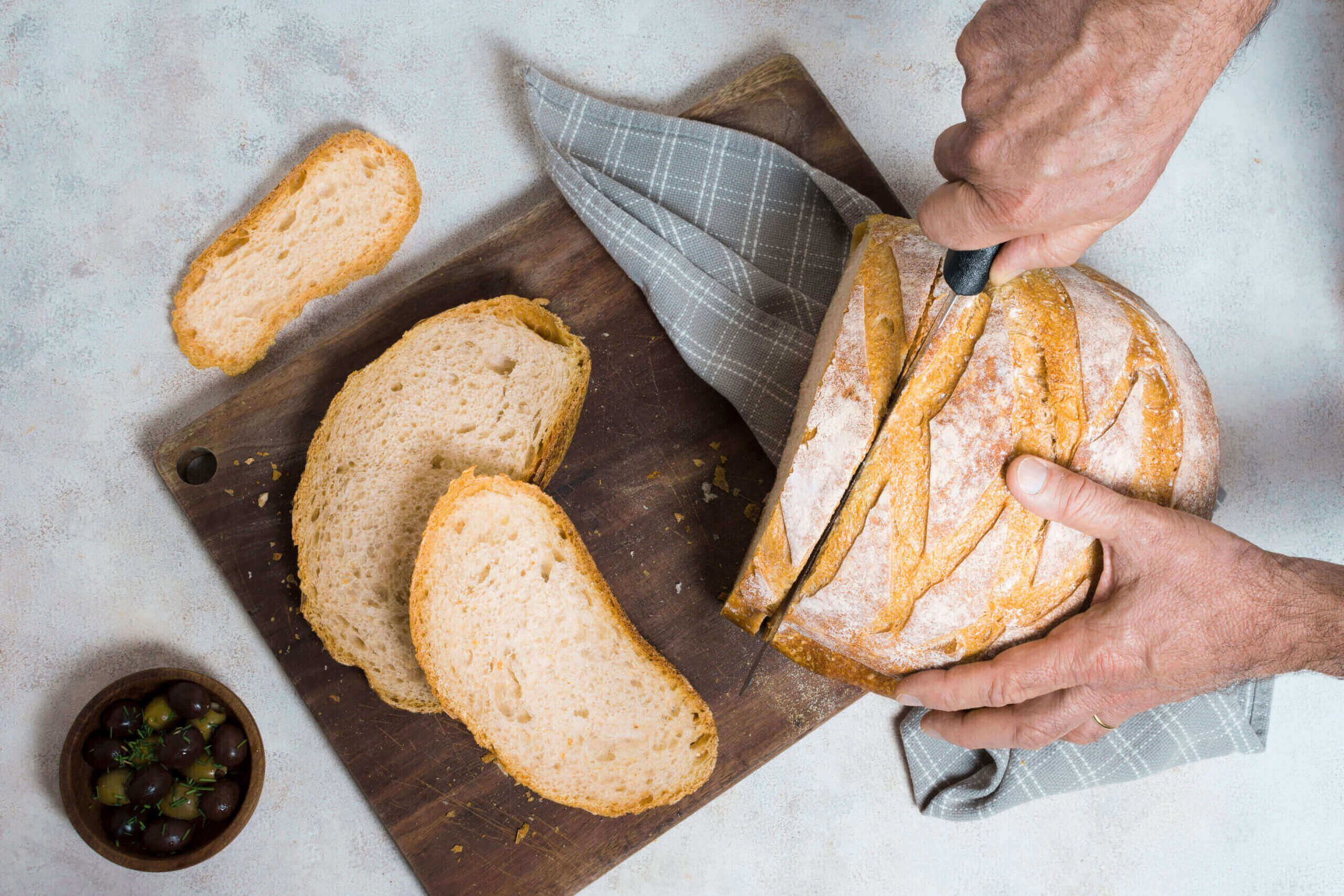 A person slices a round loaf of rustic bread on a wooden cutting board with a gray cloth. Several bread slices and a small bowl of olives are also on the board.