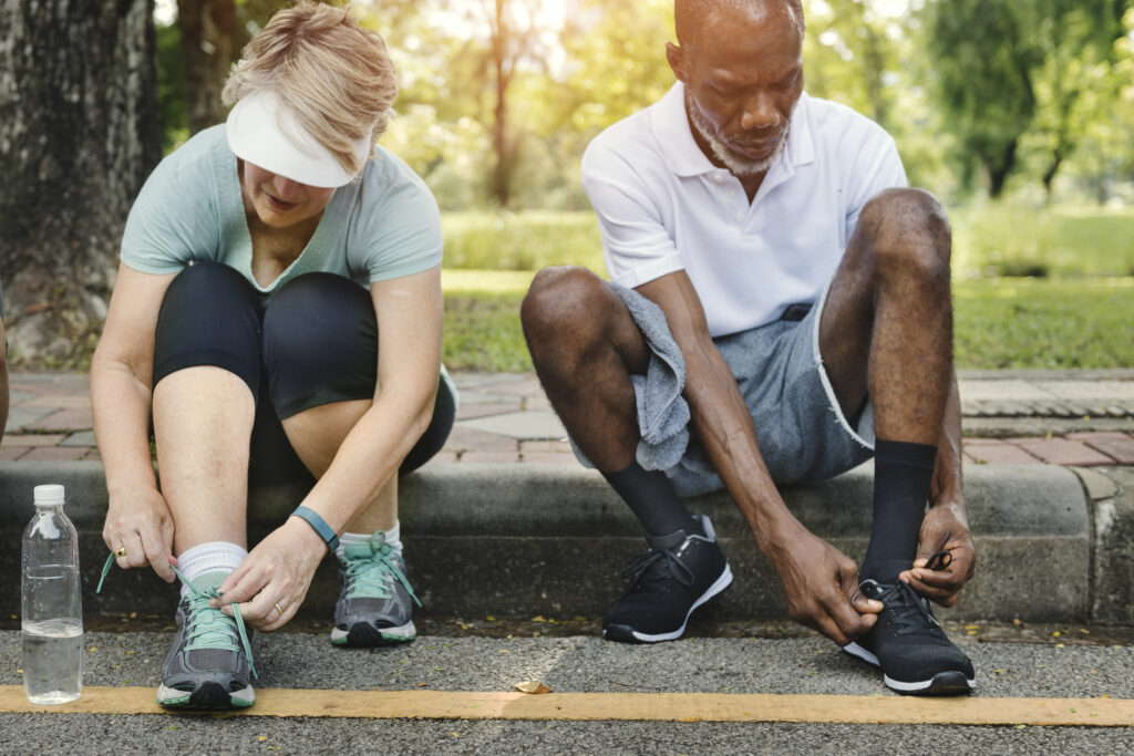 Two adults sit on a curb outdoors tying their shoelaces before exercising. The woman wears a visor and teal sneakers, while the man wears a white shirt and black sneakers. A water bottle is on the ground beside them.