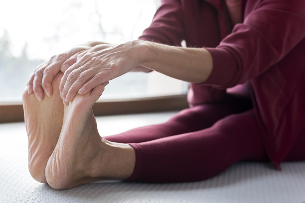 Close-up of a person in burgundy clothing sitting on the floor, stretching forward to touch their toes with both hands. The focus is on their legs, feet, and hands.