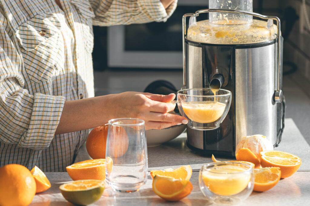 A person is juicing fresh oranges with a juicer, holding a glass cup under the spout. Sliced oranges and an empty glass sit on the kitchen counter.