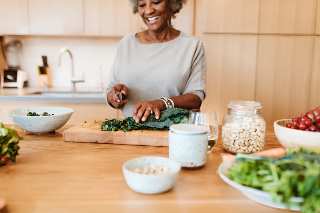 An older woman smiles while chopping leafy greens on a wooden cutting board in a modern kitchen, surrounded by bowls of fresh vegetables, nuts, and a glass of water.