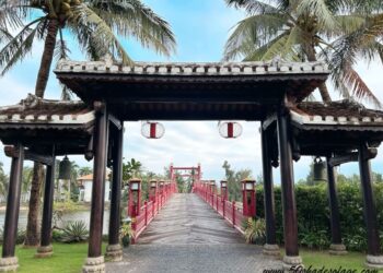 A traditional wooden entrance gate with lanterns leads to a red pedestrian bridge, surrounded by palm trees and greenery under a partly cloudy sky.