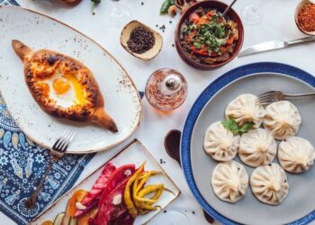 A table set with Georgian food and drink, including khachapuri with an egg, khinkali dumplings, pickled vegetables, and a bowl of stewed vegetables, arranged on patterned plates and a blue tablecloth.