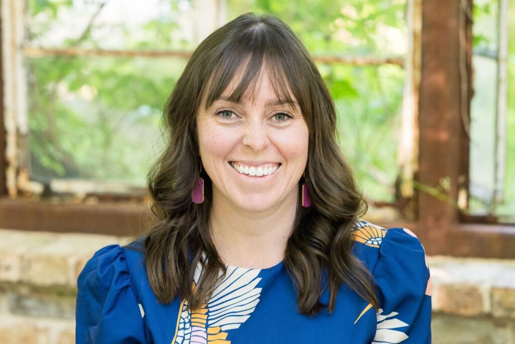 A woman with long brown hair and bangs, wearing a blue patterned blouse and pink earrings, smiles in front of a rustic window with greenery outside.