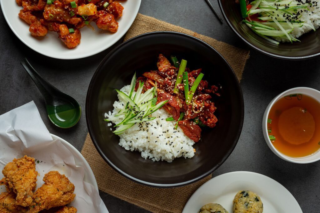 A bowl of white rice topped with sliced beef, cucumber strips, green onions, and sesame seeds sits on a table, surrounded by plates of fried chicken, saucy chicken, rice, and a cup of tea.