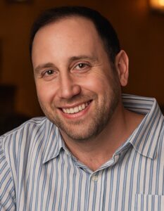 A man with short dark hair and a beard smiles warmly at the camera. He is wearing a light-colored, striped button-up shirt. The softly blurred, warmly lit background suggests he’s sharing advice on the best dog breeds for empty nesters.