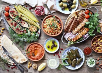 A colorful spread inspired by Turkish cuisine recipes, featuring grilled vegetables, skewered meats, dolmas, olives, nuts, flatbread, dips, rice salad, and lemon water—all beautifully arranged on a rustic wooden table.