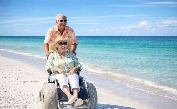 An older man smiles as he pushes an older woman in a beach-wheelchair with large wheels along the sunny, sandy shoreline. Both wear sunglasses and hats, enjoying a bright day by the turquoise ocean.