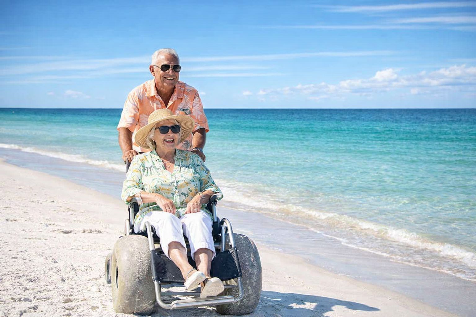 An older man smiles as he pushes an older woman in a beach-wheelchair with large wheels along the sunny, sandy shoreline. Both wear sunglasses and hats, enjoying a bright day by the turquoise ocean.