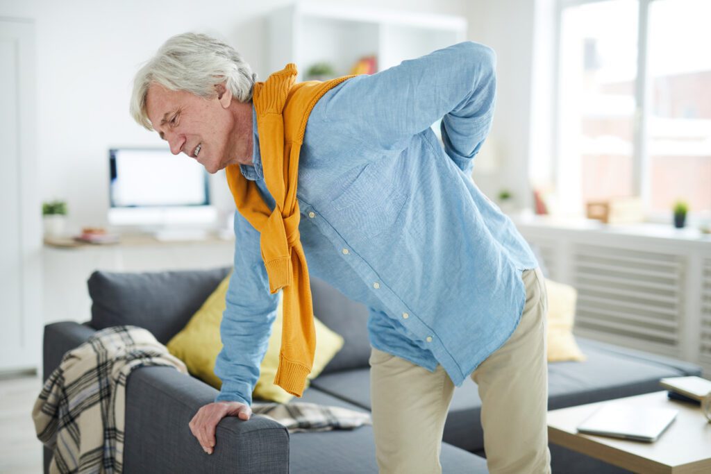 An older man with gray hair, wearing a blue shirt and yellow sweater, leans on a sofa and holds his lower back in pain inside a bright living room.