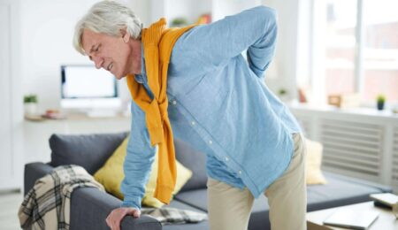 An older man with gray hair, wearing a blue shirt and yellow sweater, leans on a sofa and holds his lower back in pain inside a bright living room.