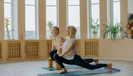 Two older adults practice yoga indoors, performing a low lunge pose on mats in a bright room with large windows and potted plants. Both look focused and relaxed.