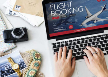 A person uses a laptop to book a flight online; the screen shows a flight booking page with a plane image. Nearby are travel essentials, suggesting planning and senior travel tips on a budget for an affordable adventure.