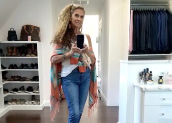 A woman with curly hair takes a mirror selfie in a walk-in closet, wearing jeans, a white shirt, and a colorful shawl. Shelves of shoes, handbags, suits, and a vanity with makeup are visible in the background.