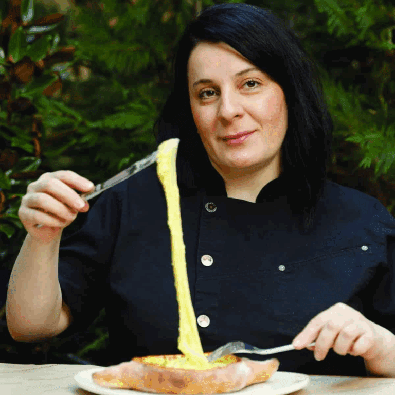 A woman with dark hair and a black chef coat smiles while pulling stretchy melted cheese from a bread boat with a knife and fork, enjoying delicious food and drink outdoors with lush greenery in the background.