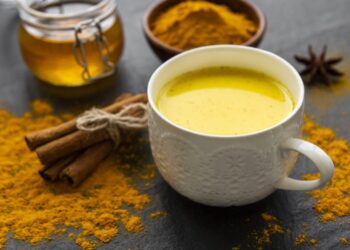 A white mug filled with golden turmeric milk sits on a dark surface, surrounded by cinnamon sticks, a jar of honey, a bowl of turmeric powder, and scattered turmeric for a vibrant, spiced presentation.