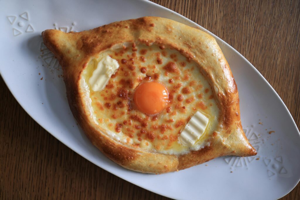 A baked Georgian cheese bread, khachapuri, filled with melted cheese, a raw egg yolk, and two small pieces of butter, served on a white oval plate atop a wooden table.