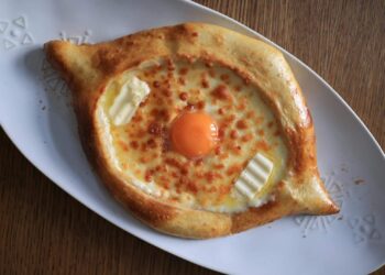 A baked Georgian cheese bread, khachapuri, filled with melted cheese, a raw egg yolk, and two small pieces of butter, served on a white oval plate atop a wooden table.