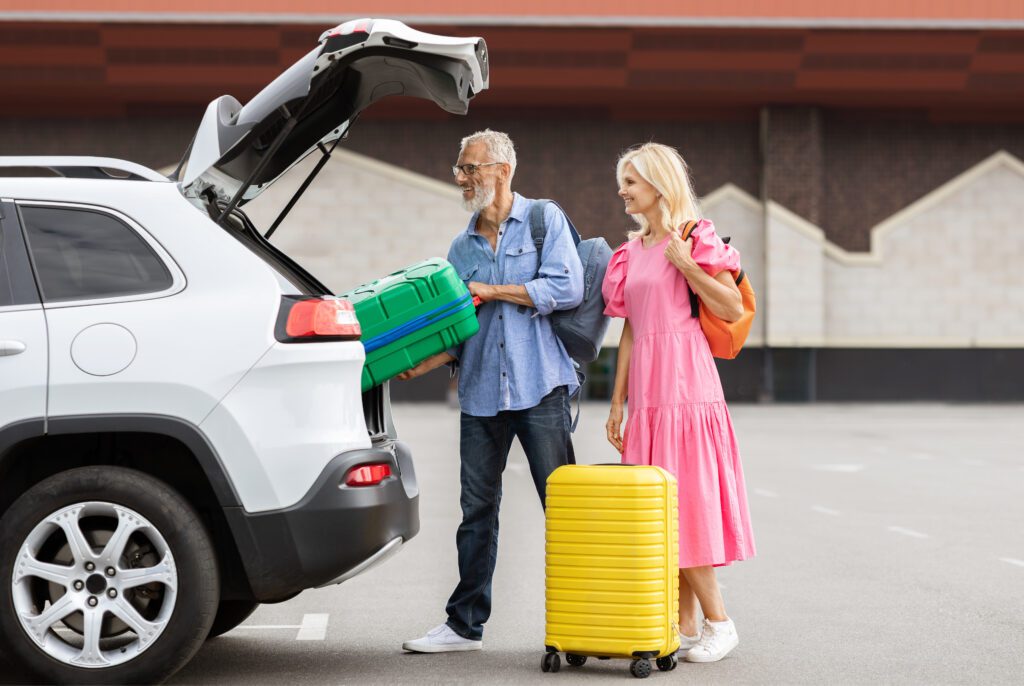An older man loads a green suitcase into the trunk of a white SUV—perhaps following senior car rental tips—while a woman in a pink dress with an orange bag and yellow suitcase stands beside him in the parking lot.