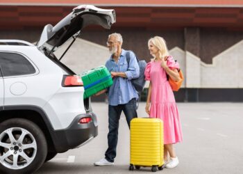An older man loads a green suitcase into the trunk of a white SUV—perhaps following senior car rental tips—while a woman in a pink dress with an orange bag and yellow suitcase stands beside him in the parking lot.