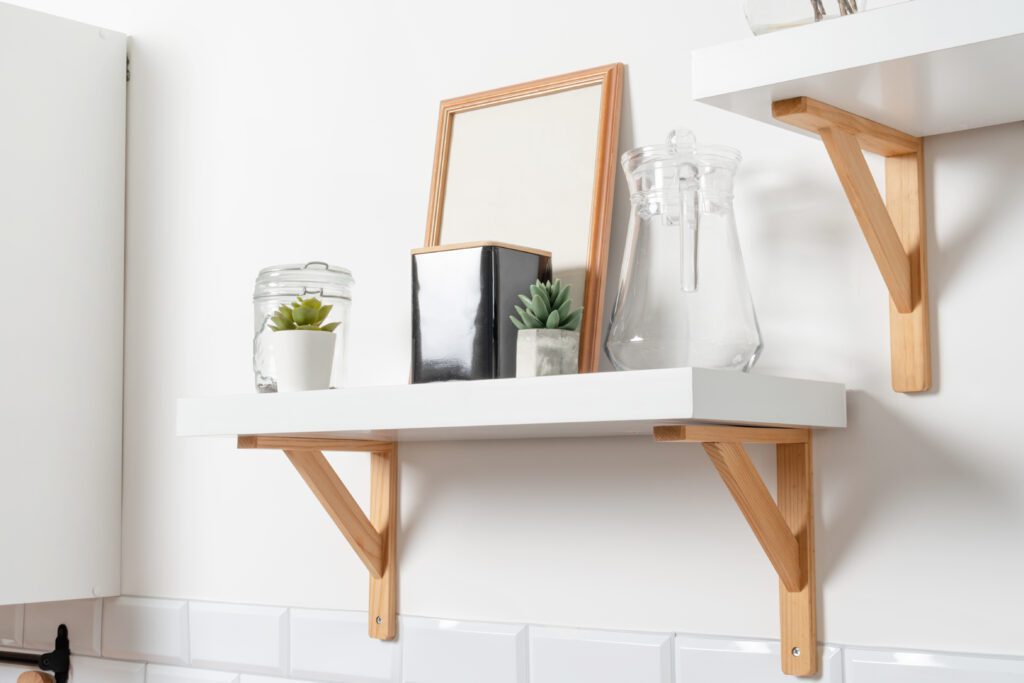 A white wall shelf with wooden brackets—perfect for budget DIY shelf makeover ideas—holds two small potted succulents, a glass jar with a lid, a glass pitcher, a framed picture, and two black books. Another shelf is partially visible above.
