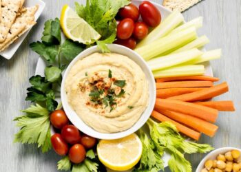 A white plate with a bowl of hummus topped with herbs and paprika, surrounded by celery sticks, carrot sticks, grape tomatoes, lemon slices, parsley, and crackers—perfect for enjoying the heart health benefits of hummus.
