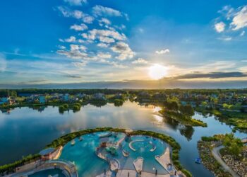 Aerial view of a large, winding swimming pool at Center Parcs Europe vacation, nestled by a calm lake at sunset, with vibrant greenery, scattered buildings, and a dramatic sky with clouds as the sun nears the horizon.