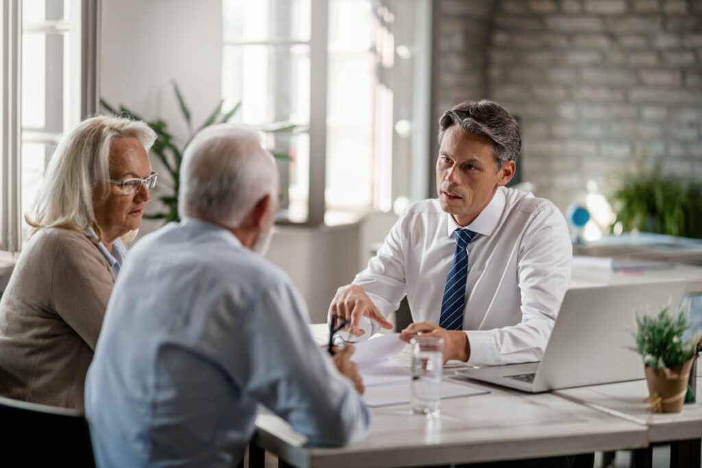 A middle-aged man in a suit discusses what a power of attorney can and can’t do with an older couple at a desk in a bright office, where a laptop, glass of water, and plant sit on the table.
