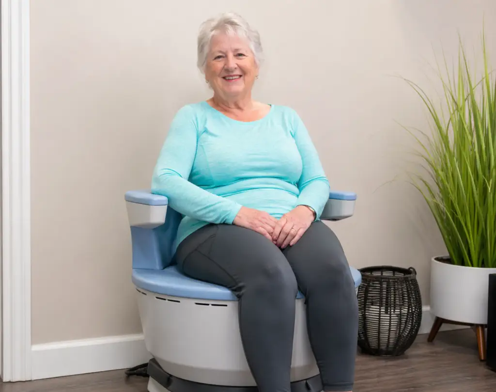 An older woman with gray hair, experiencing loss of bladder control, sits and smiles on a modern circular blue and white chair in a bright room with a plant and a basket in the background.