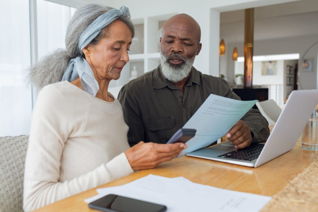 An older couple sits at a table with a laptop and documents. The woman holds a smartphone while the man reads from a sheet of paper, both appearing focused on how to maximize Social Security benefits for their retirement.