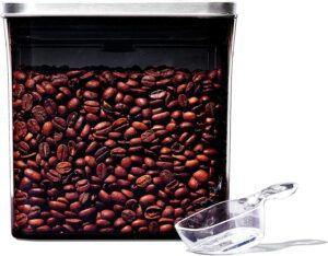 A clear square container filled with roasted coffee beans, topped with a silver lid, sits next to a transparent plastic scoop on a white background.