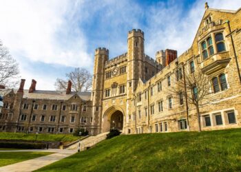 A large, historic stone building with arched entryway and tall towers stands under a blue sky in Princeton, surrounded by green grass and trees—an ideal sight to explore during a weekend in Princeton. The architecture features Gothic-style windows and detailed stonework.
