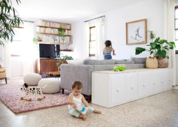 A bright living room with plants, bookshelves, and a gray couch features simple storage solutions. A toddler plays on the floor with a toy, while an older child stands on the couch as sunlight streams in through the windows.