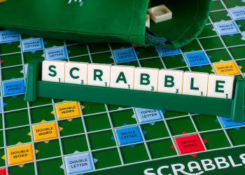 A Scrabble game board with a tile rack holding letter tiles that spell SCRABBLE. The board shows premium score squares, a green tile bag in the background, highlighting the mental benefits of Scrabble for seniors.