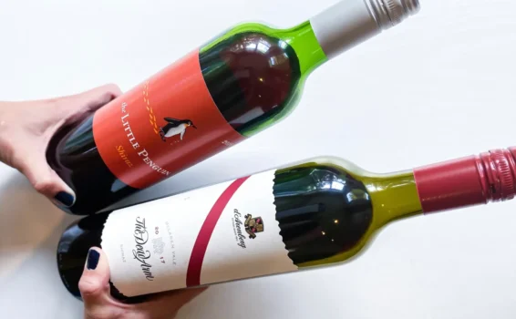 Two hands hold two bottles of red wine on a white surface. The top bottle is The Little Penguin Shiraz, featuring a playful penguin illustration; below it sits Château La Gordonne, an expensive wine with a red cap and elegant crest logo.