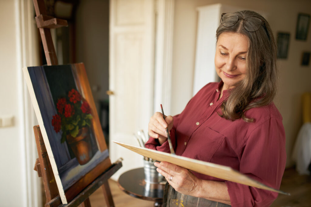 A woman with long brown hair, wearing a maroon shirt and glasses on her head, smiles while painting flowers in a pot on a canvas set on an easel in a cozy, sunlit room.