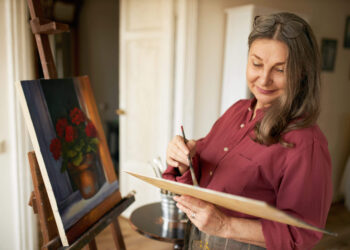 A woman with long brown hair, wearing a maroon shirt and glasses on her head, smiles while painting flowers in a pot on a canvas set on an easel in a cozy, sunlit room.