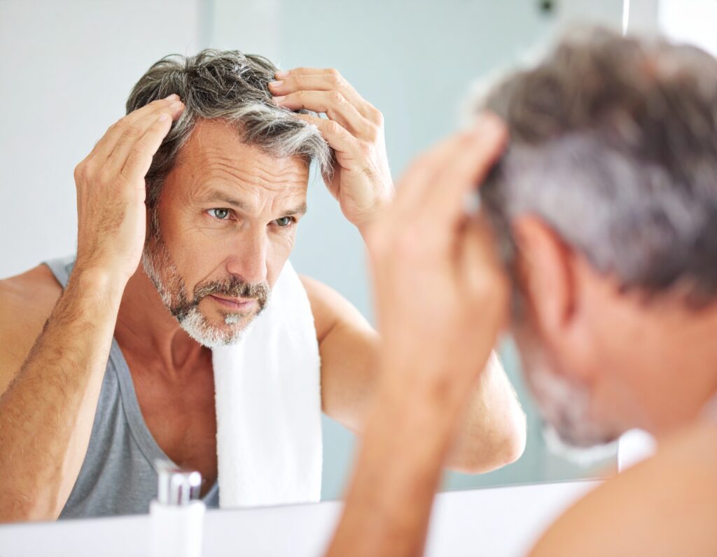 A middle-aged man with graying hair and a beard examines his scalp in the mirror, looking for hair loss warning signs as he touches his hair with both hands. He is wearing a tank top and has a towel draped over his shoulder.