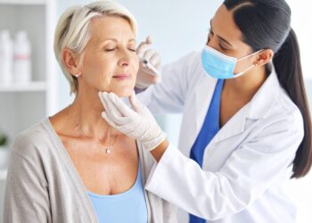 A healthcare professional wearing a mask and gloves injects a syringe into the face of an older woman with closed eyes, possibly giving a Botox after 55 cosmetic treatment, in a bright clinical setting.