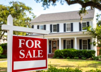 A red and white For Sale sign stands in front of a two-story white house with black shutters, a covered porch, and a well-kept lawn, surrounded by trees.