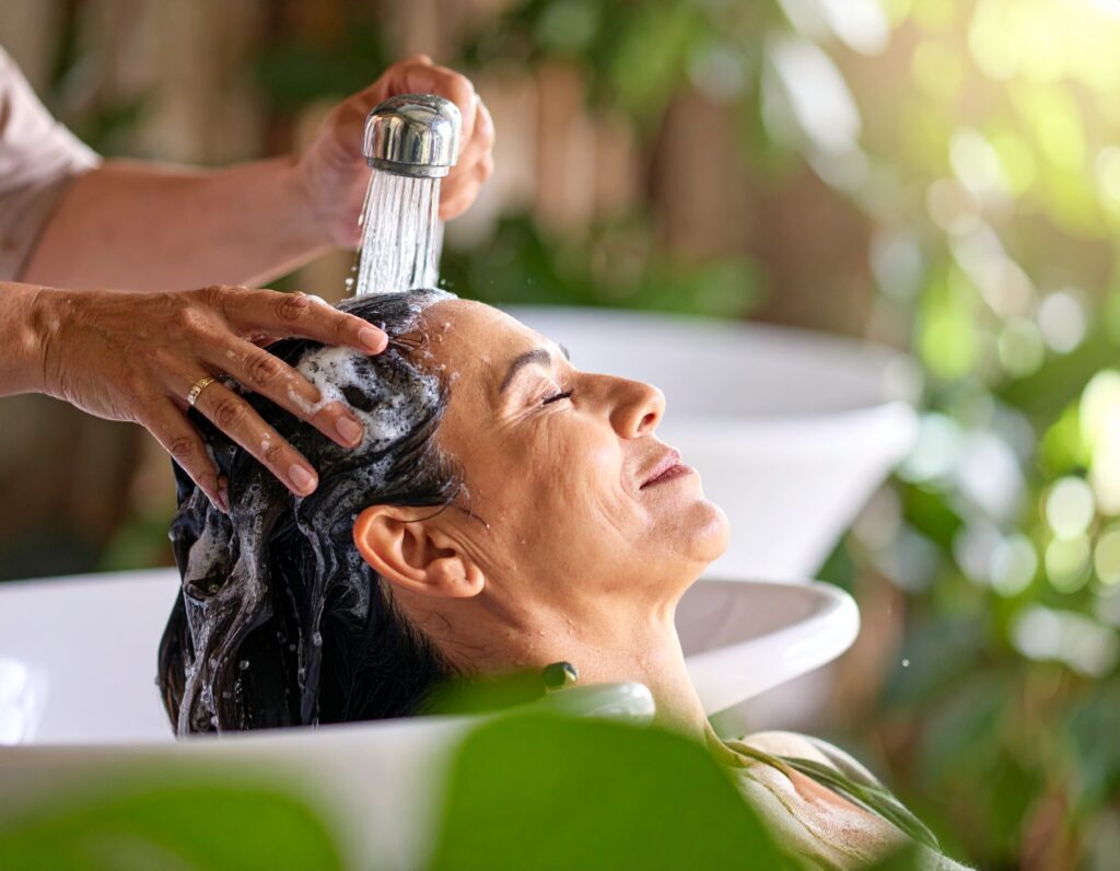 A woman enjoys natural scalp care after 55, relaxing with her eyes closed as her hair is gently washed at a bright salon surrounded by green plants.