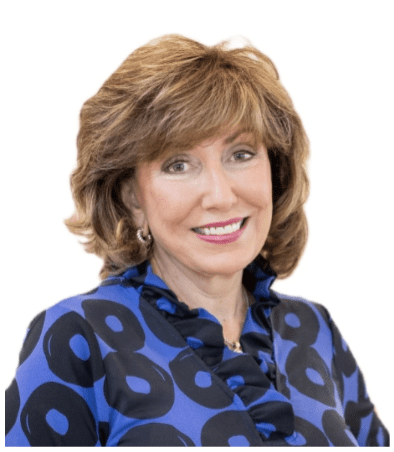 A woman with light brown, shoulder-length hair and a side part, wearing a blue blouse with a bold black circular pattern, smiles at the camera against a white background—ready for new adventures after downsizing after retirement.