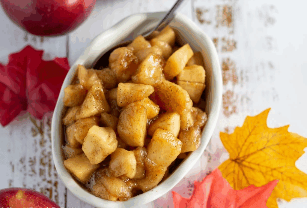 A white oval dish filled with gut-loving spiced apples sits on a rustic white wooden surface, surrounded by colorful autumn leaves. A spoon is placed inside the dish, ready to serve the cinnamon-infused goodness.