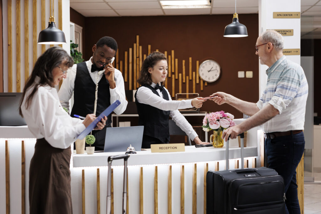 A hotel receptionist hands a key card to a guest with luggage at the front desk, offering friendly advice on trip upgrades and perks. Nearby, one staff member talks on the phone and another writes on a clipboard in the modern, welcoming setting.