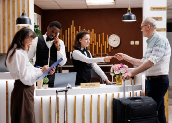 A hotel receptionist hands a key card to a guest with luggage at the front desk, offering friendly advice on trip upgrades and perks. Nearby, one staff member talks on the phone and another writes on a clipboard in the modern, welcoming setting.