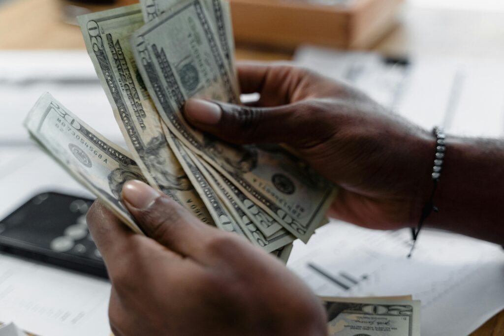 A person with dark skin counts multiple U.S. twenty-dollar bills while holding them in both hands, perhaps saving money to cut electricity costs at home, with papers and a smartphone visible in the blurred background.