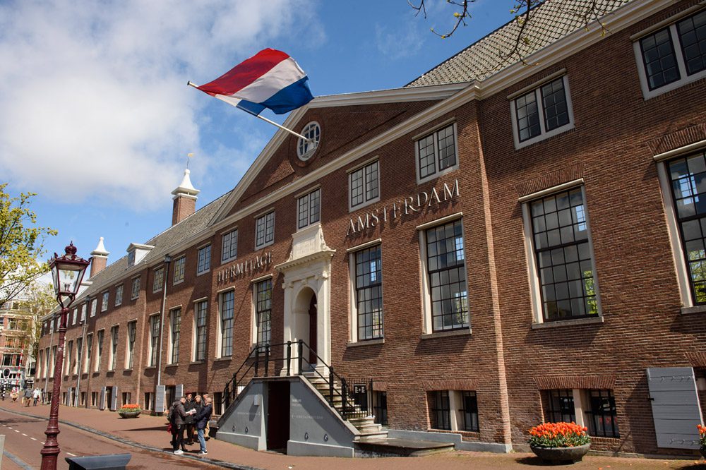 A historic brick building with Amsterdam written on the facade, large windows, and a Dutch flag flying above the entrance. On this sunny day, Amsterdam reveals its lively spirit as people gather near the front steps.