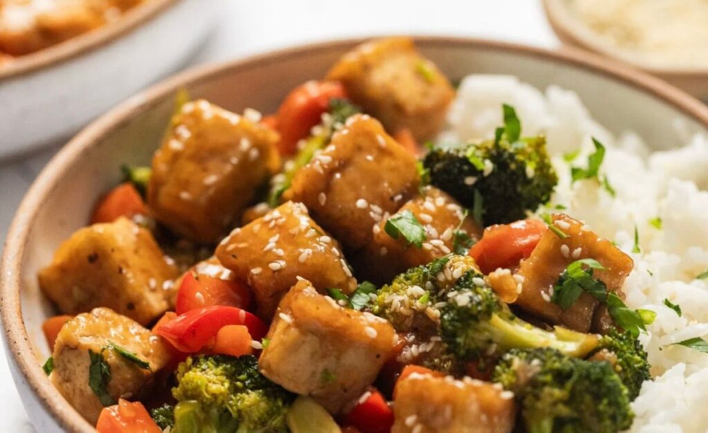 A vibrant Tofu Stir-Fry Recipe featuring stir-fried tofu, broccoli, red bell peppers, and sesame seeds, served alongside white rice and garnished with chopped fresh herbs.