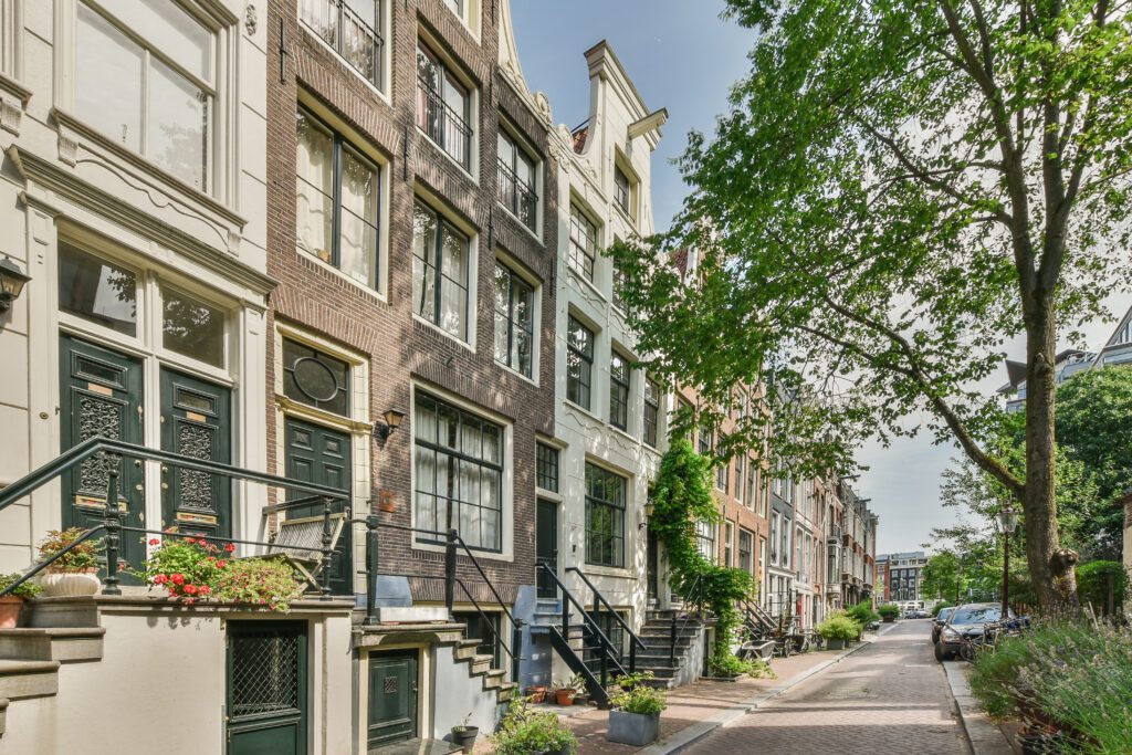 Amsterdam reveals a quiet, tree-lined street with classic tall, narrow townhouses featuring large windows and decorative doors. The sidewalk is bordered by greenery, and the scene is bright and peaceful.