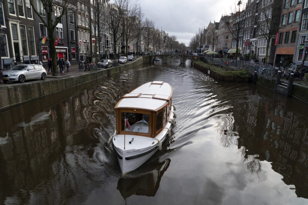 A small white boat with a wooden roof cruises down a canal as Amsterdam reveals its traditional buildings. The sky is overcast and the water ripples behind the boat, with bridges visible in the distance.